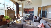 This elegantly designed single-floor community space makes an immediate impression with its dramatic, soaring ceilings and expansive floor-to-ceiling windows that flood the interior with natural light. The grand lounge area serves as the centerpiece of the space, anchored by a striking stacked-stone fireplace framed by large arched windows that offer views of the surrounding outdoors. Plush sectional sofas and wingback accent chairs are arranged around sleek glass coffee tables, creating an inviting and comfortable gathering environment. A decorative chandelier adds a refined touch overhead. Decorative columns mark the entrance foyer, which features tile flooring and leads to a reception desk area, providing a polished and professional first impression. The open-concept kitchen is equipped with dark espresso cabinetry, granite countertops, stainless steel appliances including a range and microwave, and ample storage with a full pantry — ideal for catering or community events. Warm terracotta-toned accent walls in the kitchen contrast beautifully with the neutral beige tones found throughout the rest of the space. Patterned carpeting in the lounge areas adds texture and visual interest, while tile flooring in transitional zones provides practicality. Throughout, the space balances a warm, residential ambiance with the functionality of a well-appointed communal facility.