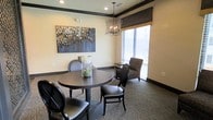 This single-floor space exudes a refined, welcoming ambiance through its thoughtful design and carefully curated details. The interior showcases warm, neutral-toned walls accented by rich dark crown molding and matching baseboards, lending the space a sense of understated elegance. Recessed lighting is complemented by a striking circular chandelier with a candelabra-style design, casting a warm glow over the room and serving as a decorative focal point. A large window fitted with horizontal blinds and a tailored Roman shade allows natural light to filter softly into the space while maintaining privacy. The seating area features a round dark-finished dining table paired with sleek, upholstered chairs in a matching dark palette, creating an intimate setting ideal for small gatherings or meetings. A plush accent chair positioned near the window adds a relaxed, conversational corner to the layout. The floors are covered in a neutral, textured carpet that adds warmth underfoot. A bold, abstract piece of wall art in muted blues and golds provides a sophisticated artistic touch. Overall, the space balances practicality with style, offering a comfortable and visually cohesive environment suitable for a variety of uses.
