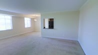 This single-floor space features an open-concept living and dining area finished with soft, neutral-toned carpeting throughout. The room is bathed in natural light from a prominently positioned window, creating a bright and airy atmosphere complemented by light green walls and crisp white trim and baseboards. A pendant light fixture hangs above the dining area, adding a subtle decorative touch. A defining architectural feature is the built-in pass-through window cut into the wall, which connects the living and dining area to the kitchen. This opening includes a dark countertop ledge and provides a clear view into the kitchen, where a black French door refrigerator and dark cabinetry are visible. The pass-through design promotes an open flow between the living spaces while maintaining a degree of separation. The overall ambiance of the space is clean, understated, and functional, with a neutral color palette that lends itself well to a variety of interior design styles. The combination of natural light, open layout, and practical architectural details makes this a versatile and welcoming living environment.
