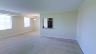 This single-floor space features an open-concept living and dining area finished with soft, neutral-toned carpeting throughout. The room is bathed in natural light from a prominently positioned window, creating a bright and airy atmosphere complemented by light green walls and crisp white trim and baseboards. A pendant light fixture hangs above the dining area, adding a subtle decorative touch. A defining architectural feature is the built-in pass-through window cut into the wall, which connects the living and dining area to the kitchen. This opening includes a dark countertop ledge and provides a clear view into the kitchen, where a black French door refrigerator and dark cabinetry are visible. The pass-through design promotes an open flow between the living spaces while maintaining a degree of separation. The overall ambiance of the space is clean, understated, and functional, with a neutral color palette that lends itself well to a variety of interior design styles. The combination of natural light, open layout, and practical architectural details makes this a versatile and welcoming living environment.