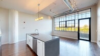 Located at 1360 Niagara St in Buffalo, NY, this one-bedroom, one-bathroom loft-style unit blends industrial character with modern finishes. The open-concept living, dining, and kitchen area serves as the heart of the home, anchored by a large kitchen island with granite-style countertops, a double stainless steel sink, white cabinetry, and stainless steel appliances. Exposed ductwork, track lighting, and oversized factory-style windows with black frames fill the space with natural light and reinforce the building's industrial heritage. Rich hardwood flooring runs throughout the entire unit. A long, well-lit hallway connects the living area to the private spaces, including the bedroom, bathroom, and multiple closets. The bedroom features a clerestory window and generous ceiling height. The bathroom is cleanly appointed with a walk-in glass shower with black framing and built-in open shelving. A pantry and in-unit laundry add practical convenience. The unit also includes access to an outdoor terrace with metal railing, offering peaceful views of the adjacent waterway and surrounding greenery — a standout feature of this Buffalo residence.