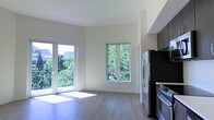 This well-designed single-floor residence offers a clean, contemporary aesthetic throughout. The open-concept living and kitchen area is flooded with natural light, courtesy of large windows and a glass-paneled balcony door that frame picturesque views of mature, lush greenery. The light-toned hardwood-style flooring runs seamlessly across the space, enhancing the airy and cohesive feel of the interior.

The kitchen is positioned along one wall and features stainless steel appliances, including a refrigerator and range, complemented by dark upper cabinetry that adds a striking contrast to the bright, neutral walls. The open layout allows the kitchen to flow naturally into the living area, making the space feel expansive and well-connected.

The living area benefits from a dual-window arrangement — a wide balcony door that opens to a private outdoor space with a metal railing, and a centered casement window that draws in additional daylight. The balcony overlooks a verdant, tree-lined setting, offering a refreshing sense of nature within an urban environment.

The overall ambiance is light, modern, and inviting, with a neutral color palette of whites and soft grays that creates a versatile backdrop. Clean lines, contemporary finishes, and an abundance of natural light define this thoughtfully laid-out space.