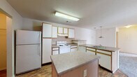 This single-floor residential unit offers a well-organized layout designed for comfortable everyday living. The kitchen serves as a functional hub, featuring white cabinetry with gold-tone trim, laminate countertops, a full suite of white appliances including a refrigerator and electric range, a double-basin sink, and a central island that provides additional prep space. Wood-look flooring runs throughout the kitchen, adding warmth to the area. A pendant light fixture transitions the space toward the open living area, which is generously sized and finished with plush carpeting. Vertical blinds covering a sliding glass door flood the room with natural light and provide direct access to the private balcony. The balcony features a wood deck, metal railing, and scenic views of mature trees and green surroundings. The bedroom is spacious and carpeted, benefiting from natural light and a ceiling fan, with a large closet offering built-in organization. The unit includes two bathrooms: one with a full tub and shower combo set on wood-look flooring, and a second with a glass-door walk-in shower, a vanity with storage, and a large mirror. Textured ceilings are consistent throughout, contributing to the unit's cohesive residential character.