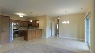 This single-story home offers an open-concept design that seamlessly connects the kitchen and living areas, creating a spacious and functional layout. The kitchen is outfitted with warm medium-toned wood cabinetry that extends to the upper walls, providing generous storage space. A mosaic tile backsplash adds texture and visual interest behind the counters, while granite countertops lend a polished, durable work surface. A center island with matching cabinetry serves as a natural focal point, offering additional prep space and casual seating potential. Pendant lighting above the island and a chandelier over the adjoining living area provide layered illumination throughout the open space. Light-toned ceramic tile flooring runs through the kitchen and main living zone, offering easy maintenance and a clean aesthetic, while a carpeted area in the living space adds warmth underfoot. Neutral beige walls complement the warm wood tones and contribute to the bright, welcoming ambiance. A hallway visible from the main living area leads to additional rooms, suggesting a well-organized floor plan that balances openness with defined private spaces.