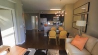 This single-floor residence offers a thoughtfully designed open-concept layout that seamlessly connects the kitchen, dining, and living areas into one cohesive and inviting space. Rich, dark hardwood floors extend throughout, grounding the space with warmth and elegance.

The kitchen is a standout feature, outfitted with dark espresso cabinetry that contrasts beautifully against stainless steel appliances, including a refrigerator, microwave, and range. A kitchen island with a stone countertop provides additional prep space and serves as a natural transition point into the dining area.

The dining space is centered around a sleek glass-top table paired with modern golden-yellow accent chairs, adding a bold pop of color to the neutral gray palette of the walls. Overhead, a striking globe chandelier with exposed Edison-style bulbs serves as a dramatic focal point, lending an upscale, contemporary feel to the space.

The living area flows naturally from the dining space, anchored by a light-toned sofa accented with a vibrant orange throw pillow and a patterned area rug. A hallway visible in the background suggests additional private rooms beyond the open living areas, completing this well-appointed and stylish residence.