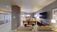 Located at 1080 Patterson St in Eugene, Oregon, this single-floor community space serves as the central hub for residents and guests alike. The layout flows seamlessly from a well-appointed lobby entrance, complete with elevator access and recessed lighting, into a series of thoughtfully designed common areas.

The main lounge area is furnished with plush, neutral-toned sofas and armchairs arranged around a wood coffee table, creating an inviting gathering space. A wall-mounted flat-screen television and coordinating media console anchor one side of the room, while floor lamps provide warm ambient lighting. Decorative artwork and fresh floral arrangements add color and character throughout.

Adjacent to the lounge, a formal dining area features a rich wood table surrounded by upholstered chairs, illuminated by a sleek pendant light fixture. A large mirror and window with natural light enhance the room's open feel.

The hallways are carpeted in a soft, textured pattern and lined with framed artwork, leading residents past a mailbox alcove and a charming decorative horse sculpture. A complimentary refreshment station outfitted with light wood cabinetry, dark granite countertops, and a coffee service offers a welcoming touch for residents and visitors. The overall ambiance is warm, calm, and residential in character.