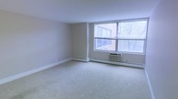 This single-floor space offers a clean and functional layout with a bright, open living area as its centerpiece. The room is finished with soft, neutral-toned carpeting and smooth walls in a warm off-white hue, creating a calm and welcoming atmosphere throughout.

Large windows dominate one wall of the living area, allowing generous amounts of natural light to fill the room while offering views of the surrounding neighborhood, including mature trees and adjacent brick architecture. The windows feature built-in blinds, providing both privacy and light control as needed.

A wall-mounted air conditioning unit and baseboard heating run along the perimeter of the space, ensuring year-round climate comfort. The simple, uncluttered design of the room provides a versatile canvas that can accommodate a variety of furniture arrangements and personal styles.

The overall ambiance is understated and practical, with an emphasis on light, openness, and ease of use. The neutral palette and carpeted flooring lend the space a quiet, residential feel, making it well-suited for those seeking a comfortable and adaptable living environment.