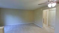 This single-floor residential space offers a clean, functional layout designed for comfortable everyday living. The main living area is generously sized, featuring wall-to-wall carpeting in a warm neutral tone that adds a soft, understated feel underfoot. The walls are finished in a muted sage-beige palette, creating a calm and cohesive atmosphere throughout. White baseboards provide a crisp contrast against the walls and carpeting, lending a tidy, well-maintained appearance to the space. A ceiling fan is mounted overhead, contributing to year-round comfort and air circulation. An electrical outlet panel is visible along one wall, and standard outlets are thoughtfully placed for practical use. The room opens through a wide, architecturally clean doorway into an adjoining area, suggesting a flowing, connected floor plan that allows for easy movement between spaces. The overall ambiance is bright and neutral, offering a versatile blank canvas well-suited to a variety of interior styles and furnishing arrangements. The simple, uncluttered design of the space makes it highly adaptable for both relaxation and daily activities.