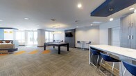 This contemporary club room is designed with both leisure and social interaction in mind. The open-concept layout is anchored by a sleek, dark-framed pool table positioned at the center of the space, illuminated by a pair of modern pendant lights that add a warm, inviting glow. A wall-mounted flat-screen TV set against a bold navy blue accent wall serves as a focal point for gathering and entertainment. The room features bar-height tables paired with modern blue chairs, providing casual seating arrangements ideal for conversation or casual dining. The neutral-toned carpet, accented with vibrant orange geometric patches, adds a playful yet polished design element to the floor. Recessed lighting throughout the ceiling ensures the space remains well-lit, complemented by large windows along one wall that invite natural light and offer views of the outdoors. The clean white walls and contemporary finishes contribute to an overall atmosphere that is modern, functional, and welcoming — a versatile amenity space well-suited for both relaxed gatherings and lively recreation.