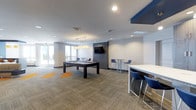 This contemporary club room is designed with both leisure and social interaction in mind. The open-concept layout is anchored by a sleek, dark-framed pool table positioned at the center of the space, illuminated by a pair of modern pendant lights that add a warm, inviting glow. A wall-mounted flat-screen TV set against a bold navy blue accent wall serves as a focal point for gathering and entertainment. The room features bar-height tables paired with modern blue chairs, providing casual seating arrangements ideal for conversation or casual dining. The neutral-toned carpet, accented with vibrant orange geometric patches, adds a playful yet polished design element to the floor. Recessed lighting throughout the ceiling ensures the space remains well-lit, complemented by large windows along one wall that invite natural light and offer views of the outdoors. The clean white walls and contemporary finishes contribute to an overall atmosphere that is modern, functional, and welcoming — a versatile amenity space well-suited for both relaxed gatherings and lively recreation.