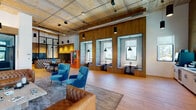 This single-floor amenity lobby combines industrial-chic architecture with warm, contemporary design to create a dynamic and welcoming communal space. Exposed concrete ceilings with visible ductwork and track lighting lend an urban loft aesthetic, while wide-plank wood flooring and warm wood-paneled accent walls add texture and warmth throughout.

The central lounge area anchors the space with a curated mix of tufted leather sofas and plush velvet armchairs in rich teal and cognac tones, arranged around a stone coffee table atop a layered area rug. A dramatic living moss wall serves as a striking focal point near the reception desk, which features glowing neon signage and a bold circular art piece.

A row of built-in banquette nooks set into a walnut wood wall provides semi-private seating for focused work or quiet conversation, each illuminated by pendant lighting and looking out through large windows to the surrounding exterior. Adjacent to this area, an enclosed workspace with glass partition walls and art deco-inspired tilework offers a more formal collaborative environment.

The mailroom area features a full bank of sleek black parcel lockers and decorative tilework, maintaining the space's design consistency. A beverage bar with a professional coffee machine, under-counter refrigeration, sink, and bright window views rounds out the amenities. Together, these elements create a sophisticated, multifunctional environment designed for both community and productivity.