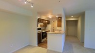 This single-floor residence at Spring House On The Park offers a thoughtfully designed open-concept layout that connects the kitchen and living areas. The kitchen is well-appointed with rich dark wood cabinetry providing ample storage, a white electric range, and a full-size refrigerator. A peninsula with a built-in sink and generous counter space serves as a natural divider between the kitchen and the adjacent living area, making it an ideal spot for casual dining or meal preparation. Track lighting throughout the space provides adjustable, focused illumination, complementing the recessed ceiling lights that brighten the hallway and entry areas. The living area features neutral-toned carpet, contributing to a warm and comfortable atmosphere, while the kitchen transitions to hardwood-style flooring for easy maintenance. The entryway, visible from the main living space, features a standard door with a straightforward, functional design. The light, neutral wall tones throughout the unit create a clean, versatile backdrop. Overall, the space offers a practical and cohesive environment well-suited for everyday living.