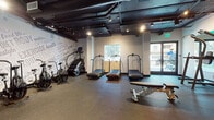 Located at 833 Fedora St in Los Angeles, this single-floor fitness facility is designed to support a comprehensive workout experience. The space features an industrial-style aesthetic, with exposed dark ceilings, visible ductwork, and track lighting that lends a modern, energetic atmosphere throughout.

The cardio area is outfitted with multiple treadmills — including curved, non-motorized models — as well as stationary bikes and a stair climber, all arranged to maximize floor space and ease of use. Natural light filters in through windows and glass entry doors, offering views of the surrounding streetscape and enhancing the overall training environment.

The strength and functional training zone houses a full rack of dumbbells, a cable machine, adjustable benches, plyo boxes, and a large wall-mounted flat-screen TV backed by floor-to-ceiling mirrors. A dedicated functional training area includes TRX suspension trainers, battle ropes, a heavy punching bag, medicine balls, and kettlebells, supporting high-intensity and cross-training workouts.

Durable rubber flooring covers the entire space, providing safety and sound absorption across all training zones. Motivational typography murals line the walls, reinforcing a culture of health and performance. The facility is thoughtfully organized to accommodate a wide range of fitness activities within a cohesive, purpose-built environment.
