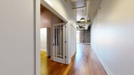 This 2-bedroom, 1-bathroom unit showcases a compelling industrial-loft aesthetic rooted in its architectural character. Warm hardwood flooring runs throughout the single-floor layout, connecting each room cohesively. Exposed brick walls are a defining feature, most prominently displayed in the combined kitchen and living area, where they stretch toward elevated ceilings fitted with visible ductwork and pendant lighting — details that underscore the unit's converted-building origins.

The living area receives generous natural light through large windows and a glass balcony door, which opens to an outdoor space with open city views. The kitchen is outfitted with rich, dark wood cabinetry, light countertops, and a full suite of stainless steel appliances including a refrigerator, range, dishwasher, and microwave.

A hallway connects the main living area to the private rooms, featuring a glass-paneled interior door and louvered closet doors. Both bedrooms offer hardwood flooring and neutral walls, with the primary bedroom providing a comfortable, well-proportioned layout. The bathroom features stone-look tile flooring, grab bars, and a combined tub and shower. A dedicated closet off the hallway provides additional storage.