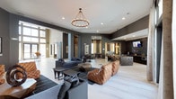 This single-floor space presents a sophisticated, open-concept layout designed for both comfort and social engagement. The expansive main area features soaring ceilings that enhance the sense of grandeur, while a striking wrought-iron ring chandelier serves as a dramatic focal point overhead. Recessed lighting complements the ambient glow throughout the space.

The lounge area is anchored by a rich, tufted cognac leather sectional paired with slate-grey sofas and geometrically shaped accent tables atop a bold area rug, creating a refined yet inviting seating arrangement. Dark charcoal accent walls contrast elegantly with the light-toned flooring, lending the space a contemporary, upscale atmosphere.

Toward the rear, a bar or refreshment counter with dark cabinetry and counter seating offers a social hub, complete with pendant lighting above and a mounted television nearby. A secondary dining or game area is visible in the background, further demonstrating the space's versatility. Large windows along the perimeter allow natural light to filter in, brightening the overall environment and connecting the interior to the outside.