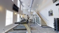 This two-story fitness center offers a spacious and well-equipped environment designed to support a comprehensive workout experience. The main floor features a row of cardio machines, including treadmills from TRUE Fitness, alongside a multi-station strength-training cable system, providing users with a versatile range of exercise options. The layout is thoughtfully organized, allowing ample space between equipment for comfortable use.

The space is defined by its dramatically high ceilings, which lend an open and airy atmosphere throughout. Exposed HVAC ductwork runs along the upper ceiling, complementing the modern industrial aesthetic, while a ceiling fan helps maintain airflow. Recessed lighting and wall-mounted sconces provide consistent, well-distributed illumination, and tall windows along the perimeter allow natural light to filter in.

A staircase with clean white railings leads to the second level, suggesting additional fitness space or amenity areas above. The flooring features a patterned, textured carpet suited for a gym environment, and a wall-mounted flat-screen television adds a functional entertainment element. The overall ambiance is clean, bright, and contemporary.