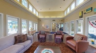 Located at 150 Arbors Cir in Gahanna, Ohio, this single-floor community space makes an immediate impression with its expansive, light-filled lounge area. The centerpiece of the space is a dramatic great room boasting soaring double-height ceilings lined with a continuous band of clerestory windows, flooding the interior with natural light and creating an open, airy atmosphere. Warm neutral tones on the walls are complemented by rich hardwood-style flooring and plush area rugs, lending the space a welcoming and refined character. The lounge is thoughtfully appointed with a mix of comfortable seating, including plush sofas, leather club chairs, and colorful ottomans arranged around a central coffee table — ideal for both relaxed gatherings and casual conversation. French doors on multiple sides of the room open to outdoor areas, seamlessly connecting the interior to the surrounding exterior. A credenza with decorative accents adds a homey touch, while recessed lighting supplements the abundant daylight. Hallways lead to additional areas beyond the main lounge, suggesting further functional spaces within the facility. Overall, the space exudes a warm, inviting ambiance designed to foster community and comfort.