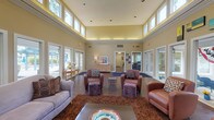 Located at 150 Arbors Cir in Gahanna, Ohio, this single-floor community space makes an immediate impression with its expansive, light-filled lounge area. The centerpiece of the space is a dramatic great room boasting soaring double-height ceilings lined with a continuous band of clerestory windows, flooding the interior with natural light and creating an open, airy atmosphere. Warm neutral tones on the walls are complemented by rich hardwood-style flooring and plush area rugs, lending the space a welcoming and refined character. The lounge is thoughtfully appointed with a mix of comfortable seating, including plush sofas, leather club chairs, and colorful ottomans arranged around a central coffee table — ideal for both relaxed gatherings and casual conversation. French doors on multiple sides of the room open to outdoor areas, seamlessly connecting the interior to the surrounding exterior. A credenza with decorative accents adds a homey touch, while recessed lighting supplements the abundant daylight. Hallways lead to additional areas beyond the main lounge, suggesting further functional spaces within the facility. Overall, the space exudes a warm, inviting ambiance designed to foster community and comfort.