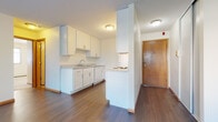 This single-floor unit offers a practical and clean layout designed for comfortable everyday living. The kitchen is arranged in a galley configuration, featuring white cabinetry with sleek bar-pull hardware, laminate countertops, a stainless steel sink, and white appliances including an electric range, refrigerator, and a range hood above. Rich, dark hardwood-style flooring runs throughout the kitchen and living areas, adding warmth to the neutral white walls. The kitchen opens toward the living space via a partial dividing wall, creating a semi-open feel while maintaining defined zones. The living area is generously sized, bathed in natural light from large windows fitted with horizontal blinds, and includes a wall-mounted air conditioning unit and baseboard heating. The bedrooms are carpeted in a neutral tone, each featuring natural light from windows with blinds and baseboard heating units. The bathroom is finished with classic white square wall tiles surrounding a full soaking tub, complemented by hardwood-style flooring. A warm-toned wood entry door completes the unit's inviting entryway. Overall, the space presents a clean, functional aesthetic with consistent finishes throughout.