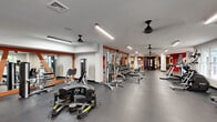 This expansive fitness center offers a generously sized, single-floor layout designed to accommodate a full spectrum of workout needs. The space is thoughtfully organized, with distinct zones for strength training, cardio, and free weights, allowing multiple users to exercise simultaneously without crowding.

The strength training area is lined with a diverse selection of cable machines, weight benches, a Smith machine, and a full dumbbell rack, all arranged along mirrored walls framed in warm orange accents that add a dynamic visual character to the space. These large mirrors serve both a functional and aesthetic purpose, amplifying the sense of openness throughout the room.

The cardio zone, situated near the entrance, benefits from natural light streaming through large windows and glass double doors, creating an inviting and airy atmosphere. A row of ellipticals and treadmills lines this section, complemented by ceiling fans that promote air circulation throughout the facility.

Durable rubber flooring covers the entire space, providing both safety and comfort underfoot. Overhead, linear LED lighting ensures the facility is consistently and evenly illuminated. A bold motivational mural reading "DON'T QUIT" anchors one end of the room against a striking steel-blue accent wall, setting an energizing tone. Framed artwork is also displayed throughout, adding a polished, welcoming finish to the overall environment.