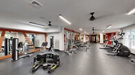 This expansive fitness center offers a generously sized, single-floor layout designed to accommodate a full spectrum of workout needs. The space is thoughtfully organized, with distinct zones for strength training, cardio, and free weights, allowing multiple users to exercise simultaneously without crowding.

The strength training area is lined with a diverse selection of cable machines, weight benches, a Smith machine, and a full dumbbell rack, all arranged along mirrored walls framed in warm orange accents that add a dynamic visual character to the space. These large mirrors serve both a functional and aesthetic purpose, amplifying the sense of openness throughout the room.

The cardio zone, situated near the entrance, benefits from natural light streaming through large windows and glass double doors, creating an inviting and airy atmosphere. A row of ellipticals and treadmills lines this section, complemented by ceiling fans that promote air circulation throughout the facility.

Durable rubber flooring covers the entire space, providing both safety and comfort underfoot. Overhead, linear LED lighting ensures the facility is consistently and evenly illuminated. A bold motivational mural reading "DON'T QUIT" anchors one end of the room against a striking steel-blue accent wall, setting an energizing tone. Framed artwork is also displayed throughout, adding a polished, welcoming finish to the overall environment.