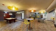 This dynamic amenity floor is designed for recreation, fitness, and social engagement, offering a diverse range of activity zones within a single, well-appointed level. The centerpiece game room is defined by its textured brick-style accent walls, bold geometric-patterned carpeting, and eye-catching pendant lights in yellow and orange hues. It features a pool table, ping-pong table, and high-top bistro seating, creating an energetic yet comfortable gathering space. Adjacent to it, a cozy lounge nook provides a more relaxed atmosphere with plush leather seating, rich burnt-orange accent walls, and framed billiard-themed artwork. The dedicated fitness center is equipped with a multi-station weight machine, free weights, and wall-to-wall mirrors that amplify the sense of space, all under recessed lighting with decorative etched-glass door panels. A separate exercise studio offers an open, flexible layout with a ceiling fan, rubber flooring, and a full rack of fitness equipment including stability balls and medicine balls. The expansive open activity hall stands out with its vivid lime-green epoxy floors, whitewashed brick walls, a dark exposed ceiling, and colorful mural accents, making it ideal for a variety of uses. Transitional corridors feature warm orange walls, whitewashed brick, and intimate seating areas, tying the entire floor together with a cohesive, lively aesthetic.