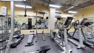 This fitness center is a comprehensively outfitted exercise facility designed to support a full range of workout routines. The space features warm yellow walls, dark rubber flooring, and an open ceiling with exposed infrastructure and fluorescent lighting that creates a bright, energetic atmosphere throughout.

The cardio zone is lined with multiple treadmills and elliptical machines from Life Fitness, accompanied by wall-mounted televisions for entertainment during workouts. A water cooler station is conveniently positioned nearby to keep users hydrated.

The strength training area is equally well-appointed, featuring a Life Fitness cable machine, a pull-up and dip station, adjustable benches, and a dedicated free weights section with a full rack of dumbbells in graduated sizes. Large floor-to-ceiling mirrors span multiple walls, amplifying the sense of space and allowing users to monitor their form during exercises.

The layout is thoughtfully organized to separate cardio and strength zones while maintaining an open, accessible flow between them. A ceiling fan contributes to air circulation, and posted informational signage adds a practical touch. The facility is compact yet efficiently arranged, making it a functional and well-rounded space for daily fitness needs.