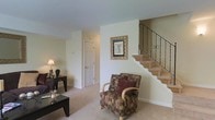 This two-floor residence offers a functional and comfortable layout highlighted by a ground-level living area that sets a warm, inviting tone throughout the space. The living room features wall-to-wall carpeting in a neutral tone, providing a soft and cohesive foundation that complements the light, cream-colored walls. A carpeted staircase with a decorative wrought-iron railing rises from the main living area, serving as both a practical and visually appealing architectural element connecting the two floors. The living space is sized to accommodate seating arrangements comfortably, as evidenced by the patterned armchair and accompanying dark wood coffee and side tables. A framed floral artwork on the wall adds a touch of character to the space. The main level also includes an interior door, suggesting additional rooms or storage accessible from the living area. Natural light filters into the space, contributing to the overall bright and airy atmosphere. The neutral color palette and simple finishes throughout create a clean, understated aesthetic that offers a versatile backdrop for personalization.