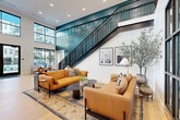 Located at 110 E 36th Street in Charlotte, NC, this thoughtfully designed space makes a bold architectural statement from the moment you enter. The open-plan layout seamlessly connects the kitchen and living room, creating a fluid, welcoming environment ideal for gathering and entertaining.

The living area is anchored by dramatic double-height ceilings that amplify the sense of openness, while expansive floor-to-ceiling windows along multiple walls flood the interior with natural light and frame views of the surrounding outdoor courtyard. A striking staircase with teal-accented railings adds a sculptural focal point and reinforces the vertical drama of the space. Warm natural hardwood flooring runs throughout, providing a cohesive foundation beneath thoughtfully arranged seating areas furnished with rich cognac leather sofas and a sculptural curved sectional.

The kitchen is a standout feature, showcasing deep teal cabinetry paired with a large waterfall-edge marble island offering generous bar seating. Earthy tile backsplash, open shelving with decorative lighting, and stainless steel appliances complete a kitchen that is as functional as it is visually refined. Tall potted olive trees positioned throughout the living area introduce organic texture and reinforce the connection between the interior and the lush outdoor spaces visible beyond the glass.