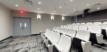 This private screening room delivers a polished, cinema-quality experience within a sleek, contemporary design. Multiple rows of white leather theater seats are arranged in a tiered layout, each equipped with built-in cup holders and dark armrests, ensuring comfortable viewing from every angle. The room's neutral color palette — featuring warm gray textured wall treatments, dark baseboard trim, and a boldly patterned gray carpet — creates a sophisticated yet immersive atmosphere.

At the front of the room, a large projection screen is anchored above a built-in media cabinet finished with a warm wood-tone countertop and dark paneled storage below, providing a refined focal point. A ceiling-mounted projector is positioned toward the rear of the space, paired with wall-mounted speakers to deliver a comprehensive audio-visual setup. Recessed ceiling lighting supplements sleek wall sconces that cast a soft, ambient glow along the side walls. A rear dividing rail separates the seating rows, adding visual structure to the tiered arrangement. Double exit doors are clearly marked, and the overall layout prioritizes both functionality and an elevated viewing experience.