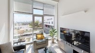 This contemporary single-floor unit showcases a thoughtfully designed layout that balances shared living with private accommodations. The open living area is a standout, boasting soaring ceilings and expansive floor-to-ceiling windows that flood the space with natural light and frame impressive city skyline views. The living room comfortably accommodates seating and entertainment areas, creating an inviting communal atmosphere.

The kitchen is well-appointed with light wood cabinetry extending to the ceiling, stainless steel appliances including a refrigerator, range, and microwave, and a dining table with bar-style seating for casual meals. Light wood-tone flooring carries throughout the common areas, adding warmth to the modern aesthetic.

Multiple private bedrooms each feature their own floor-to-ceiling windows with city views, high ceilings, built-in wardrobe storage, and dedicated desk space — ideal for study or work. The bedrooms offer carpeted flooring for comfort. Bathrooms are clean and functional, featuring wood-toned vanities with white countertops, large mirrors, and shower areas with ample storage. A shared hallway connects the private spaces efficiently. Overall, the unit delivers a bright, urban living experience with a cohesive modern design throughout.