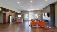 This contemporary single-floor clubhouse is designed to serve as a dynamic communal gathering space. The layout flows seamlessly across several distinct areas, each thoughtfully furnished and finished with modern design sensibilities.

The main lounge is a generously proportioned open-concept area anchored by bold orange modular seating arranged over a vivid geometric-patterned area rug. A wall-mounted flat-screen TV and a sleek fireplace are set into a charcoal accent wall, creating a focal point for relaxation and socializing. Large glass doors along the perimeter fill the space with natural light and provide views of an outdoor pool area.

Adjacent to the lounge, the fully equipped kitchen features warm wood-tone cabinetry, stainless steel appliances including a full-size refrigerator, white countertops, and a striking multicolored mosaic tile backsplash that adds a vibrant artistic touch. A kitchen island with orange bar stools extends the entertaining functionality of the space.

A secondary media room offers a quieter retreat, highlighted by a built-in wall unit with recessed display niches, decorative greenery, and an embedded television beneath an arched architectural detail.

The entry hall welcomes visitors with rich hardwood-style flooring, arched transom windows, and an open, airy layout accented with teal seating and a tall potted plant. Recessed lighting throughout maintains a warm, inviting ambiance across all areas.