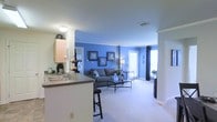 Located at 650 Bentley Ridge Boulevard in Lancaster, Pennsylvania, this single-floor residence offers a thoughtfully designed open-concept layout that seamlessly connects the kitchen and living areas. The kitchen features light wood cabinetry, a dark countertop with a built-in sink, and a breakfast bar with seating — ideal for casual dining or entertaining. It opens directly into the expansive living room, which is defined by a bold blue accent wall adorned with framed artwork, adding a vibrant yet cohesive character to the space. The living area is finished with plush wall-to-wall carpeting and is furnished with a comfortable gray sofa and a glass-top coffee table, creating a warm and inviting atmosphere. Natural light flows in generously through large windows dressed with blinds and floor-length dark curtains, brightening the room throughout the day. Crown molding along the ceiling adds a refined architectural touch, while neutral white walls and trim throughout provide a clean, polished backdrop. A hallway visible from the main living space suggests additional rooms beyond, rounding out this well-designed, move-in-ready home.