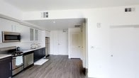 This single-floor space showcases a clean, contemporary design defined by crisp white walls and rich dark hardwood-style flooring that runs continuously throughout the unit. The open-concept layout connects the kitchen to the main living area, creating a seamless flow between spaces. The kitchen is outfitted with dark cabinetry contrasted by upper cabinets with glass-panel doors, a stainless steel refrigerator, and a sink with modern fixtures. Recessed lighting in the kitchen area provides focused illumination, complementing the natural brightness reflected off the white walls. A step-up threshold subtly separates the kitchen from the adjoining living space, adding a mild architectural distinction. Toward the rear of the unit, two white doors are visible — one being a solid entry door and the other featuring louvered panels, likely leading to a closet or utility space. HVAC vents are integrated into the ceiling, ensuring climate control throughout. The overall ambiance is minimal and modern, offering a neutral, move-in-ready environment with a functional layout well-suited for everyday living.