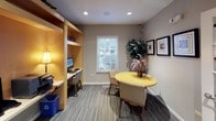 This single-floor amenity space is thoughtfully designed to offer residents a variety of functional areas in a cohesive, professional environment. The business center is a standout feature, comprising built-in workstations finished in light maple wood with integrated shelving above, providing a clean and organized workspace for focused productivity. Recessed lighting and a warm lamp illuminate the area, creating an inviting atmosphere for work or study.

Adjacent to the workstations, a casual meeting nook features a round yellow accent table paired with upholstered chairs, ideal for informal gatherings or collaborative sessions. A potted plant and framed wall art add a welcoming, lived-in quality to the space, while a window with blinds allows natural light to filter in.

The space transitions seamlessly through classic white French doors with glass pane detailing, lending an air of elegance and openness throughout. The flooring shifts between patterned carpet and light-toned tile, subtly defining each functional zone. Framed artwork adorns the neutral gray walls, contributing to a polished aesthetic.

Beyond the French doors, a hallway leads to a fitness facility, visible through glass-paneled partitions, completing this well-rounded amenity offering. The overall ambiance is professional yet comfortable, striking a balance between utility and style.