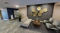 This single-floor space makes a strong first impression with its thoughtfully curated lounge area, blending contemporary design with refined decorative touches. The focal point of the seating area is a striking feature wall adorned with a bold geometric patterned wallcovering in deep navy and gold tones, against which a cluster of oversized metallic gold disc sculptures are artfully arranged — creating a dramatic and eye-catching display. A tufted light grey sofa anchors the space, paired with a rich walnut-toned coffee table and a complementary armchair, all set atop a layered grey area rug that adds texture and warmth underfoot. A spiral gold tabletop accent piece adds an additional layer of curated detail to the arrangement. The flooring throughout features a muted, multi-toned carpet that contributes to the space's understated elegance. Recessed ceiling lighting provides a clean, modern illumination across the room. A lush potted tropical plant near the corridor entrance introduces a natural, refreshing element that softens the space's polished aesthetic. The overall ambiance is one of sophisticated comfort, making the space well-suited for welcoming guests or facilitating relaxed, professional gatherings.