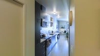 This single-floor space showcases a thoughtfully designed open layout where the kitchen flows directly into the living area. The galley-style kitchen is outfitted with dark, contemporary cabinetry that extends floor to ceiling, providing ample storage. Stainless steel appliances, including a built-in microwave, oven, and dishwasher, are seamlessly integrated into the design. Open shelving along the upper wall adds visual interest and additional display space, while the light-toned countertops create a striking contrast against the dark cabinetry. Light wood-style flooring runs throughout, contributing to a clean, modern aesthetic. Recessed and flush-mount ceiling lighting illuminates the kitchen corridor efficiently. Beyond the kitchen, the living area benefits from natural light filtering through a window, creating a warm and inviting atmosphere. A dining area with seating is visible, making good use of the available space. The overall design balances functionality with a modern, urban aesthetic, making efficient use of the linear floor plan.