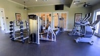 This fitness center is a thoughtfully designed, single-floor space built to support a comprehensive strength and conditioning routine. The room is outfitted with professional-grade Precor strength machines, including a multi-station cable system, a weight rack, and an ab station, all arranged to maximize usability and flow throughout the space. The durable rubber flooring provides both safety and comfort underfoot, making it well-suited for a variety of exercises. Overhead, a ceiling fan ensures steady air circulation, contributing to a comfortable workout environment. A wall-mounted flat-screen television is positioned for easy viewing from multiple areas of the room, adding an entertainment element to training sessions. The space is finished with clean white trim and sage-green walls, giving it a polished yet functional aesthetic. A set of elegant French doors along the back wall opens to an adjacent warmly lit lounge or clubroom, visible through the glass panes, adding an inviting sense of depth and connectivity to the overall facility. Recessed lighting throughout keeps the space bright and well-illuminated. The overall ambiance strikes a balance between a professional gym environment and a refined, community-oriented setting.