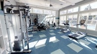 This single-story fitness center is designed to provide a comfortable and functional workout environment. The space is bathed in natural light, courtesy of large windows lining multiple walls, creating an open and energizing atmosphere. A ceiling fan overhead aids in air circulation, contributing to a pleasant exercise experience.

The flooring consists of a patterned blue and gray modular carpet tile layout, offering both durability and a clean aesthetic. The cardio zone is well-represented with elliptical trainers, a stationary bike, and a treadmill positioned to take advantage of the abundant natural light and exterior views. On the strength training side, a cable machine and free weight area with barbells and weight plates provide options for resistance-based workouts.

A standout feature is the motivational typographic accent wall, displaying fitness-related words and phrases that add energy and personality to the space. The overall ambiance is bright, modern, and welcoming, making it a functional and visually appealing facility suited for a range of fitness routines.