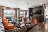 This well-appointed single-floor space is designed to accommodate a variety of social functions, combining a lounge area, dining space, and fully equipped kitchen within an open, flowing layout. Rich wood-look tile flooring runs throughout, anchoring the contemporary aesthetic and providing visual continuity between zones.

The lounge area is a focal point of the space, centered around a bold dark accent wall clad in horizontal shiplap paneling. A wall-mounted flat-screen television is paired with a dark wood media console, while colorful framed artwork adds visual interest to either side. Cognac leather armchairs and a large tufted sectional sofa in charcoal gray are arranged for comfortable gathering, accented with vibrant orange throw pillows that echo the warm tones of the rust-colored drapery throughout.

The yellow-painted lounge wall adds an energetic contrast, with coordinating artwork and wall-mounted swing-arm sconce lighting enhancing the layered ambiance. Large windows and glass doors invite abundant natural light and provide views of the outdoor surroundings.

The kitchen is well-equipped with dark cabinetry, granite countertops, a tile backsplash, stainless steel appliances, and decorative pendant lighting over an island. A dining nook with a wood table, bench seating, and large windows completes the space, offering a bright setting for casual meals.