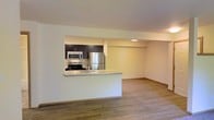 This single-floor space offers a clean, modern design centered around an open-concept layout that seamlessly connects the kitchen and living area. The kitchen is a standout feature, showcasing dark espresso cabinetry that contrasts beautifully against light-toned walls and countertops. Stainless steel appliances — including a refrigerator, range, and over-the-range microwave — lend a contemporary, polished look. A peninsula counter serves as a natural divider between the kitchen and the open living space, offering both functionality and a sense of flow throughout the area. Recessed lighting in the kitchen provides focused illumination, complementing the warm, ambient lighting of the adjoining space. The living area is generously proportioned, with light wood-toned flooring that extends throughout, adding warmth and a sense of continuity to the space. Neutral wall tones enhance the bright, airy atmosphere. Clean lines, minimal detailing, and a well-considered layout make this space feel inviting and practical, well-suited for everyday living and entertaining alike.