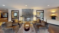 The Fireside Lounge is a thoughtfully designed social space that seamlessly blends comfort and elegance across a single floor. The centerpiece of the lounge is a dramatic floor-to-ceiling stone fireplace, crafted from natural stacked stone, which anchors the room and creates an inviting focal point. Flanking the fireplace are plush, earth-toned armchairs arranged around a round coffee table atop a textured area rug, forming an intimate seating vignette ideal for relaxed conversation.

Rich, dark hardwood flooring runs throughout the space, complemented by deep wood-trimmed windows and door frames that lend a refined, cohesive aesthetic. Large framed windows and mirrors are strategically placed to amplify natural light and create a sense of openness. Wall-mounted sconces cast a warm, ambient glow throughout, enhancing the lounge's welcoming atmosphere.

A dedicated dining and café-style seating area features bistro-height tables paired with sleek black leather bar chairs positioned along windows overlooking snow-dusted greenery and stone exterior architectural elements. Additional dining seating with dark chairs provides further flexible gathering options. Recessed ceiling lighting and crown molding add polished architectural detail. The overall ambiance is warm, upscale, and conducive to both casual socializing and quiet relaxation.