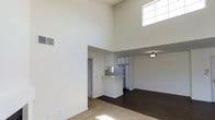 This single-floor space makes an immediate impression with its dramatically high ceilings that lend an expansive, airy quality to the main living area. Large clerestory windows positioned near the ceiling top flood the room with natural light, enhancing the sense of openness and volume throughout the space. The living area features light-toned carpet flooring that transitions into rich, dark hardwood in the adjacent areas, creating a clear and attractive delineation between zones. The open-plan kitchen is thoughtfully integrated into the layout, featuring white cabinetry that extends to a generous height, a refrigerator, and a peninsula-style breakfast bar that serves as both a functional workspace and a casual dining spot. The kitchen's placement allows for easy interaction with the surrounding living and dining areas, making the overall layout ideal for everyday living. Neutral, warm wall tones throughout the space contribute to a clean and inviting ambiance. A flush-mount ceiling light fixture in the adjoining area provides additional illumination, complementing the abundant natural light. Doorways leading to other areas of the home are visible, suggesting a well-connected and practical floor plan. Overall, the space combines modern functionality with an open, light-filled atmosphere.