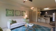 This single-floor residence offers a thoughtfully designed open-concept layout that seamlessly connects the living area to the kitchen, creating a sense of spaciousness and easy flow. The living room serves as the heart of the home, featuring a ceiling fan overhead and neutral-toned walls that provide a calm, welcoming backdrop. A glass-top coffee table and a bold teal area rug anchor the space, adding color and visual interest against the light tile flooring. The room is accented with vibrant green decorative wall art, lending a fresh, lively character to the interior. Adjacent to the living area, the kitchen features dark cabinetry, open shelving with pendant lighting, and a breakfast bar with counter-height seating — ideal for casual dining or entertaining. The consistent use of warm neutral tones paired with green and teal accents throughout gives the space a cohesive and inviting ambiance. A louvered interior door adds a subtle architectural detail, while the overall layout emphasizes both functionality and comfort. This residence is well-suited for those seeking a modern, move-in-ready home with a stylish yet practical design.