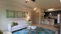 This single-floor residence offers a thoughtfully designed open-concept layout that seamlessly connects the living area to the kitchen, creating a sense of spaciousness and easy flow. The living room serves as the heart of the home, featuring a ceiling fan overhead and neutral-toned walls that provide a calm, welcoming backdrop. A glass-top coffee table and a bold teal area rug anchor the space, adding color and visual interest against the light tile flooring. The room is accented with vibrant green decorative wall art, lending a fresh, lively character to the interior. Adjacent to the living area, the kitchen features dark cabinetry, open shelving with pendant lighting, and a breakfast bar with counter-height seating — ideal for casual dining or entertaining. The consistent use of warm neutral tones paired with green and teal accents throughout gives the space a cohesive and inviting ambiance. A louvered interior door adds a subtle architectural detail, while the overall layout emphasizes both functionality and comfort. This residence is well-suited for those seeking a modern, move-in-ready home with a stylish yet practical design.