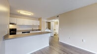 Located at 4850 50th St W in Bradenton, Florida, this single-story unit offers a practical and well-organized layout designed for comfortable everyday living. Wood-look flooring runs consistently throughout the space, providing a cohesive and easy-to-maintain surface. Neutral warm beige wall tones and textured ceilings contribute to a clean, understated ambiance.

The kitchen is positioned along one side of the open living area, defined by a peninsula counter with laminate countertops that doubles as a casual breakfast bar. White cabinetry offers ample storage overhead, while black appliances—including a refrigerator, electric range, and dishwasher—provide functional cooking amenities. The kitchen opens directly to the living area, creating a connected, airy feel.

The living area is generously proportioned, with an entry closet conveniently located near the front door. A separate bedroom features a large window that invites natural light and greenery views, complemented by a ceiling fan with light fixture.

A full bathroom includes a tub-shower combination with tiled surround. Notably, an in-unit laundry closet houses a full-size washer and dryer, a practical convenience tucked neatly behind louvered doors adjacent to the bathroom.