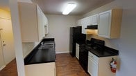 This single-floor residence offers a well-organized layout designed for comfortable everyday living. The kitchen is a standout feature, showcasing a classic galley configuration that maximizes efficiency. White raised-panel cabinetry lines both sides of the kitchen, providing generous storage space, while dark countertops offer a striking contrast that adds visual depth. The kitchen is equipped with black appliances, including a full-size refrigerator, an electric range with coil burners, and a built-in range hood, all complementing the bold, two-tone aesthetic. A stainless steel sink with a single-handle faucet is positioned along the countertop, adding a practical touch. The flooring throughout the kitchen consists of wood-style planks in a warm, medium-brown tone, lending warmth and durability to the space. A textured ceiling with flush-mounted fluorescent lighting ensures the kitchen is well-lit. The neutral wall tones keep the space feeling open and clean, while the overall design balances functionality with a timeless, understated style that suits a range of interior preferences.