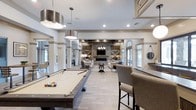 This single-floor amenity space blends modern farmhouse design with upscale comfort, creating an inviting environment perfect for socializing and leisure. The open-concept layout flows seamlessly from a billiards area to a sophisticated lounge, offering a variety of ways to enjoy the space.

At the heart of the room, a full-size pool table anchors the entertainment zone, illuminated by a large industrial-style drum pendant light. Adjacent to the billiards area is a bar-height seating section, ideal for casual gathering or spectating.

The lounge area centers around a striking floor-to-ceiling stone fireplace fitted with a mounted television, flanked by rustic barn-style wooden doors that add warmth and character. Exposed wooden ceiling beams and a chandelier overhead enhance the farmhouse aesthetic while maintaining an elevated, refined feel.

Comfortable seating arrangements with plush sofas and armchairs invite relaxation, while large arched windows along the perimeter allow abundant natural light to fill the space. Light-toned tile flooring runs throughout, complementing the neutral palette of whites, grays, and natural wood tones. Structural columns with clean architectural detailing define zones within the open floor plan without interrupting the sense of spaciousness.