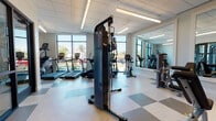 Located at 1648 Eighth Ave in Greeley, Colorado, this single-floor fitness center is thoughtfully designed to support a comprehensive workout experience. The space is organized into distinct functional zones, beginning with a cardio area positioned along a wall of large, floor-to-ceiling windows that flood the room with natural light and offer open views of the surrounding landscape. This section houses a lineup of True-brand treadmills, ellipticals, and upright bikes, all oriented to take advantage of the outdoor views.

The strength training area is equipped with a cable machine system, weight benches, a dumbbell rack, and various selectorized resistance machines, providing options for both free weight and machine-based training. A large wall mirror spans a significant portion of this zone, enhancing the sense of openness and allowing users to monitor their form during exercise.

A standout design element is a bold, full-wall black-and-white vintage photographic mural depicting athletic and motorsport scenes, adding a dynamic visual character to the space. The flooring throughout features a neutral checkered tile pattern that complements the clean, modern aesthetic. Additional amenities include a wall-mounted flat-screen television, a water fountain station, and recessed linear lighting that ensures consistent, well-distributed illumination across the entire facility.