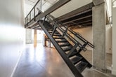 This two-floor industrial loft offers a compelling blend of raw architectural character and modern finishes. The main level showcases polished concrete floors, exposed steel beams, corrugated metal ceilings, and concrete columns that define the open layout. A well-appointed kitchen features light maple cabinetry, dark granite countertops, stainless steel appliances including a refrigerator, range, microwave, and dishwasher, complemented by large windows that invite natural light into the space. A dramatic black steel staircase with wire mesh railings serves as both a functional and visual centerpiece, connecting the two floors with industrial flair. The upper level offers an open mezzanine-style layout with concrete ceilings, steel mesh railing overlooking the main floor, and generous open space. Both bathrooms are finished with striking dark slate-tiled walls. One features a tub-shower combination, while the other offers a walk-in shower with a built-in bench and rainfall showerhead, both paired with maple vanities and granite countertops. Throughout, the interplay of raw concrete, exposed structural elements, and refined modern finishes creates a distinctive, loft-style ambiance suited for a variety of uses.